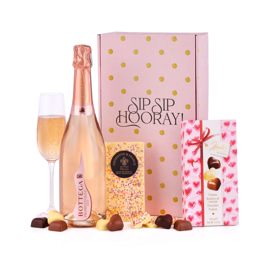 Sip Sip Hooray Prosecco Gift Box with White Chocolate bar and Pralines. The prosecco gift box reads "Sip Sip Hooray!" with golden dots covering the large prosecco gift box itself. A white bar of chocolate is leant against the gift box, with a box of personal chocolate pralines on the other side of the prosecco gift box. A large bottle of Bottega Italian Prosecco is on the left side of the image, alongside a glass of the Rose prosecco.