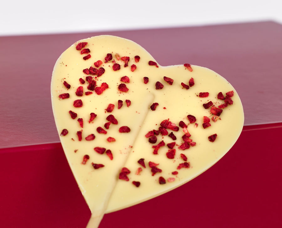 A white chocolate heart on a stick, topped with raspberry pieces, laid out in front of the alcohol free gift box, Made With Love.